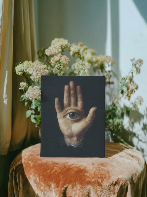 A high-contrast matte canvas print featuring a surrealist artwork. Centered against a dark, textured charcoal background, a realistic, pale hand is held up, palm forward. Embedded in the center of the palm is a detailed, light blue human eye, looking directly at the viewer. The hand emerges from a formal, dark suit cuff with a white ruffled sleeve, adding a touch of vintage, academic style. The overall mood is mysterious, gothic, and scholarly, suitable for dark academia or maximalist decor.